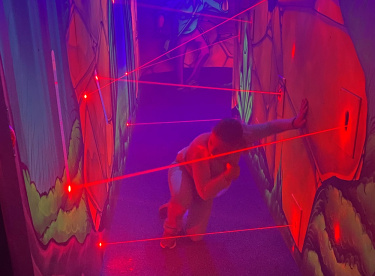 Kind bewegt sich vorsichtig durch einen schmalen, neonbeleuchteten Gang mit roten Laserstrahlen