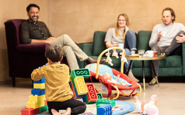 Kind spielt mit Legosteinen in der Spiellounge und die Eltern bzw. Erwachsene sitzen im Hintergrund auf dem Sofa