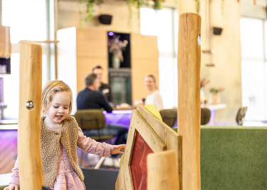 Kleines Mädchen spielt im Indoor-Spielbereich, die Eltern im Hintergrund