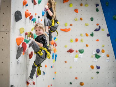 Zwei Kinder mit Klettergurten steigen an einer bunten Kletterwand nach oben 